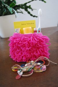 Pinkes Wolltäschchen mit Schlaufenmuster, darin eine Tube, eine Zahncreme, Parfum sowie ein Taschenbuchausgabe mit Anton Tschechovs "der Kirschgarten".
